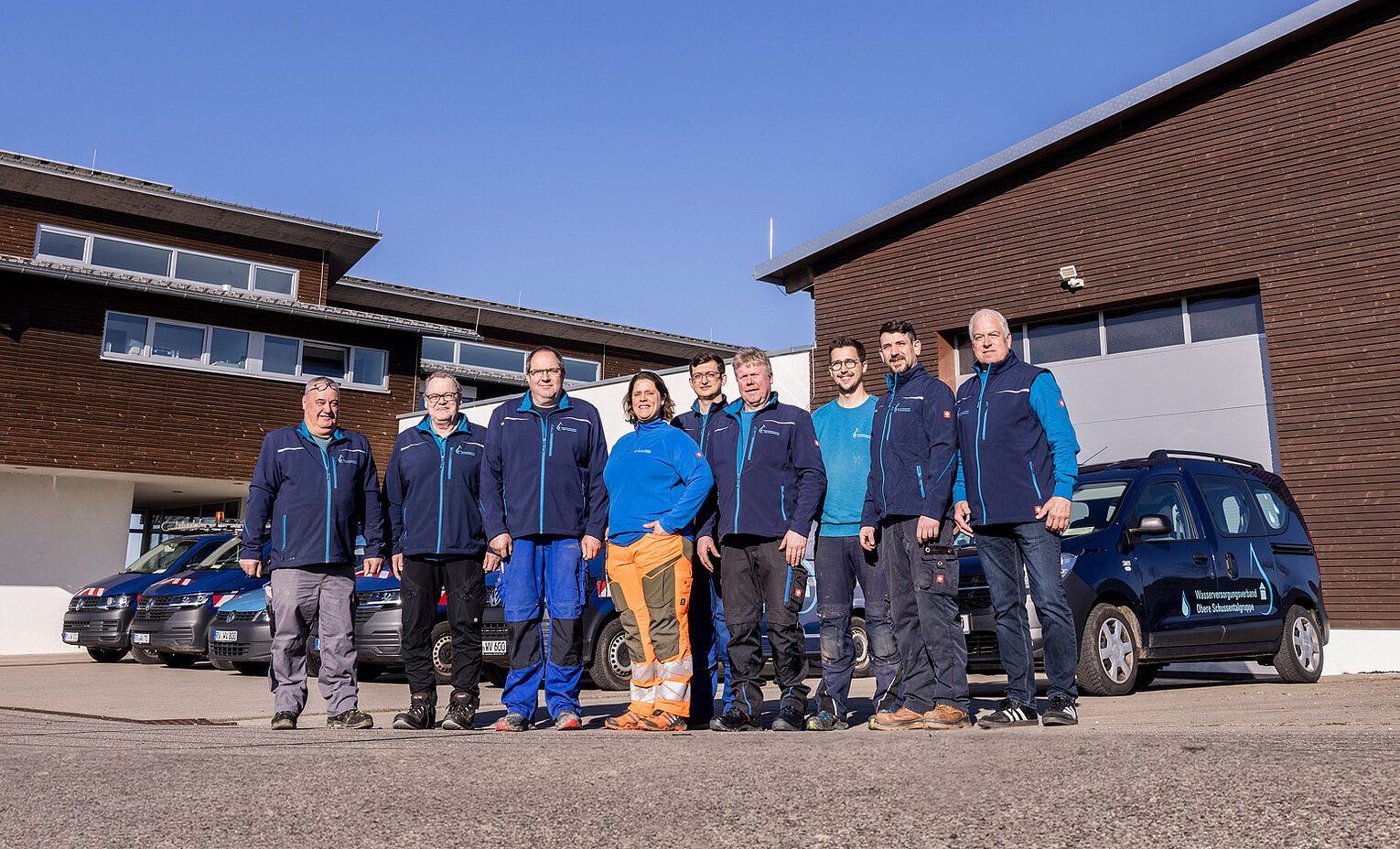 Team mit Fuhrpark Mitarbeitende des Wasserversorgungsverbands vor Einsatzfahrzeugen und Gebäuden